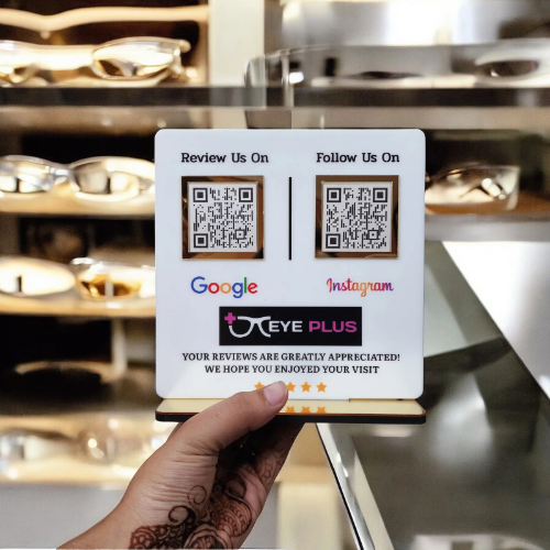 A hand with henna design holds a small white sign for "Eye Plus" featuring QR codes for Google reviews and Instagram, displayed in an optical store.