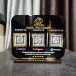 Black acrylic table stand for "Kitchen Hotel Restaurant" featuring three gold-framed QR codes for Google reviews, Instagram follows, and WhatsApp chat, resting on a dark surface.