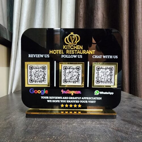 Black acrylic table stand for "Kitchen Hotel Restaurant" featuring three gold-framed QR codes for Google reviews, Instagram follows, and WhatsApp chat, resting on a dark surface.