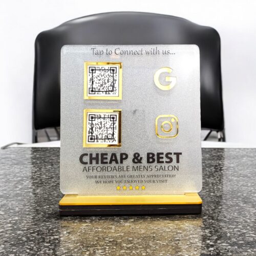 A clear acrylic sign for Cheap & Best Affordable Mens Salon displays two QR codes, a gold Google icon, and a gold Instagram icon for customer connection and reviews.